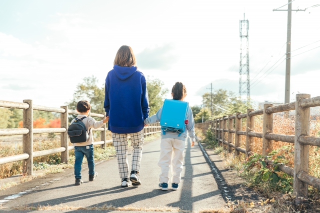 小学生と幼児の子どもを連れて歩く母親。母子家庭の母と子が下校の帰り道を歩く後ろ姿。