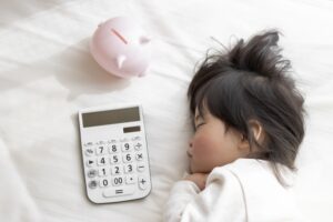 母子家庭の家計や補助金の不安を抱えながらも、子どもの生活を守ろうとする家庭のイメージ写真。電卓と貯金箱のそばで安心して眠る子ども。